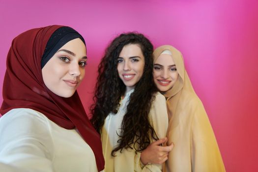 Muslim women taking selfie by mobile phone isolated on pink background. Group of pretty Arab girls in modern fashionable dress using technology