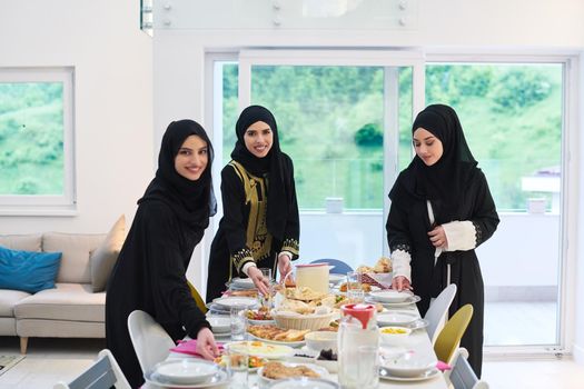 Young muslim women preparing food for iftar during Ramadan. Arabic girls in traditional abaya dresses serving table for family dinner.