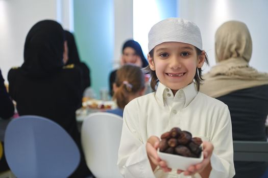 Arabian kid in the traditional clothes during iftar. Portrait of happy young muslim boy holding bowl of dates while rest of the family are eating