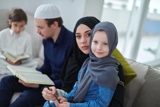 Young muslim family reading Quran during Ramadan. Parents and kids worshiping to God, in islamic clothes at modern home