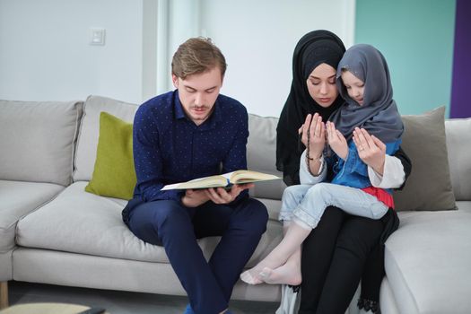Young muslim family reading Quran during Ramadan. Parents and daughter worshiping to God, in islamic clothes at modern home