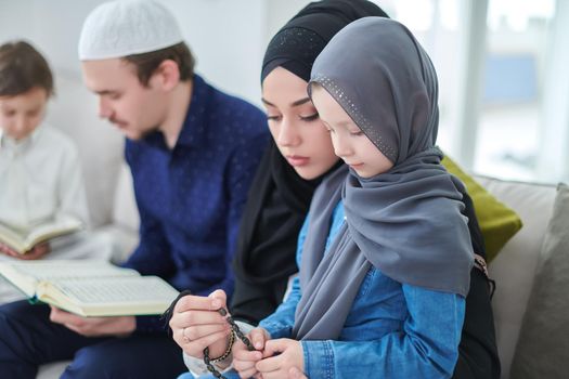 Young muslim family reading Quran during Ramadan. Parents and kids worshiping to God, in islamic clothes at modern home