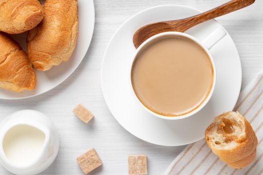 Coffee with milk in a white cup, buns and brown sugar on a white wooden table, top view, flat lay.
