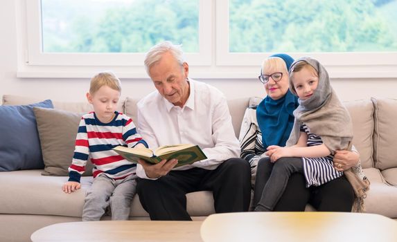 modern muslim family grandparents with grandchildren reading Quran and praying together on the sofa before iftar dinner during a ramadan feast at home