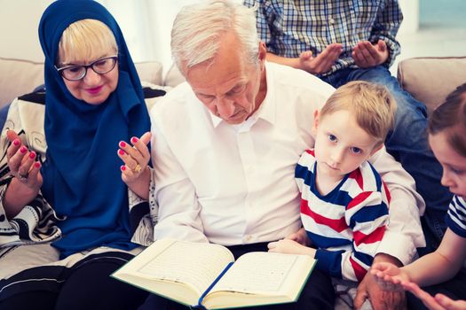 modern muslim family grandparents with grandchildren reading Quran and praying together on the sofa before iftar dinner during a ramadan feast at home