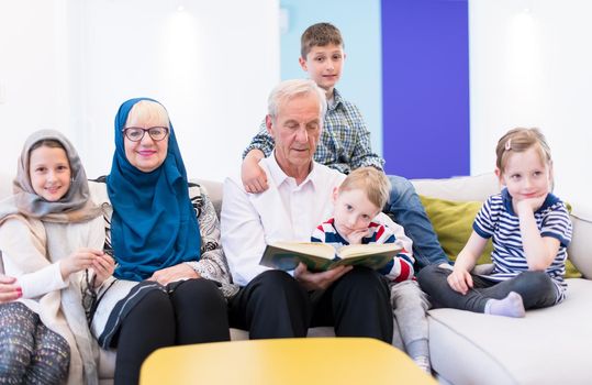 modern muslim family grandparents with grandchildren reading Quran and praying together on the sofa before iftar dinner during a ramadan feast at home