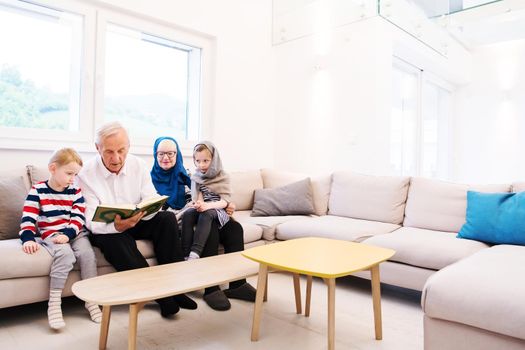 modern muslim family grandparents with grandchildren reading Quran and praying together on the sofa before iftar dinner during a ramadan feast at home