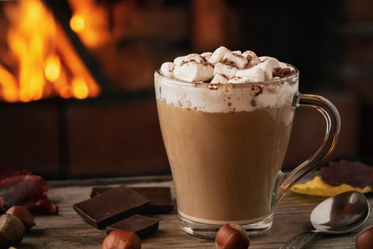 Cocoa with marshmallows and chocolate in a glass mug on a wooden table near a burning fireplace.