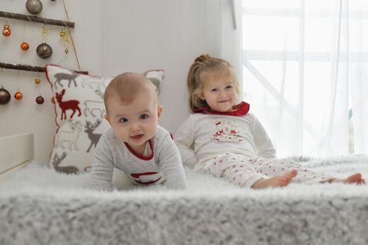 Brother and sister in Christmas pajamas are sitting on the bed with decorations.