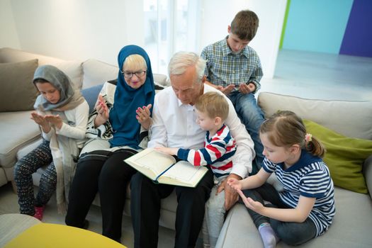 modern muslim family grandparents with grandchildren reading Quran and praying together on the sofa before iftar dinner during a ramadan feast at home