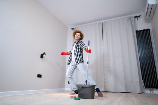 Happy mature woman mops the floor and dances and sings in a new unfurnished apartment before moving to a new house. Housewife enjoying domestic chores, doing home cleanup creatively.