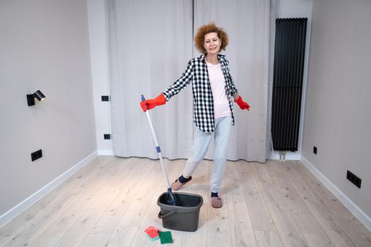 Jolly excited mature woman enjoying cleaning house, she dancing while washing floor. Happy elderly woman enjoying cleaning floors before moving to new apartment. Housework and housekeeping concept.