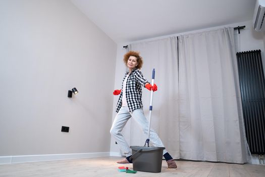 People, housework and housekeeping concept. Happy senior woman in protective gloves cleans floor and dances at home in empty apartment before moving to new home. Excited woman enjoying housekeeping.