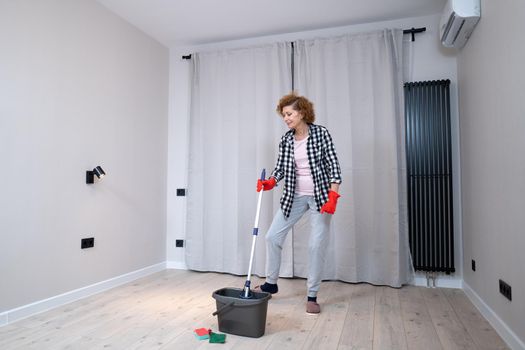 Jolly excited mature woman enjoying cleaning house, she dancing while washing floor. Happy elderly woman enjoying cleaning floors before moving to new apartment. Housework and housekeeping concept.