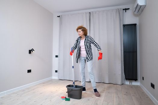 People, housework and housekeeping concept. Happy senior woman in protective gloves cleans floor and dances at home in empty apartment before moving to new home. Excited woman enjoying housekeeping.