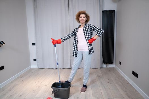 People, housework and housekeeping concept. Happy senior woman in protective gloves cleans floor and dances at home in empty apartment before moving to new home. Excited woman enjoying housekeeping.