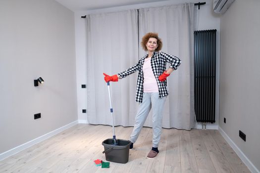 Happy mature woman mops the floor and dances and sings in a new unfurnished apartment before moving to a new house. Housewife enjoying domestic chores, doing home cleanup creatively.