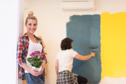 Happy couple doing home renovations, the man is painting the room and the woman hold the pot with flowers