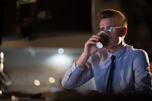 Tired businessman working late doing overtime in office at night drinking coffee to go on.