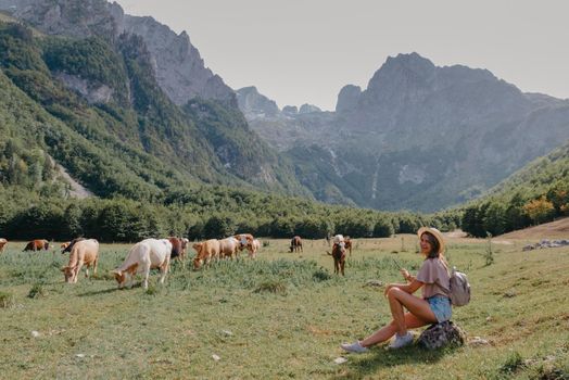 Travel, Lifestyle Concept. Beautiful woman enjoys views of the alpine village in the Alps mountains. Young woman sitting and relaxing on alpine mountain looking the cow eating a grass in the summer. Happy tourist girl traveling to Europe.