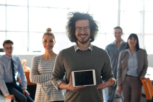 Portrait of a young businessman holding tablet in bright office with colleagues in the background