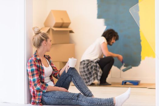 Happy couple doing home renovations, the man is painting the room and the woman is relaxing on the floor and drinking coffee