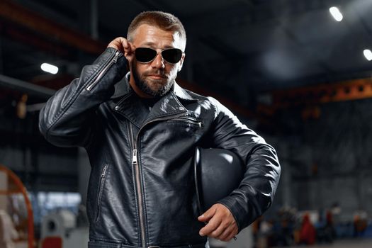 Portrait of bearded man motocyclist in dark sunglasses on dark building background