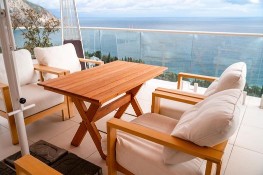 Restaurant with sea view. You can see rocks in the sea. Wooden tables and chairs with white cushions. Overcast, cloudy weather