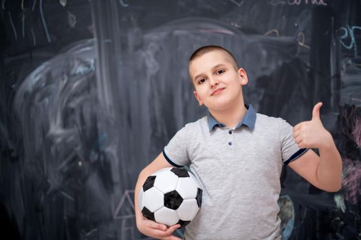 portrait of happy cute boy having fun holding a soccer ball while standing in front of black chalkboard
