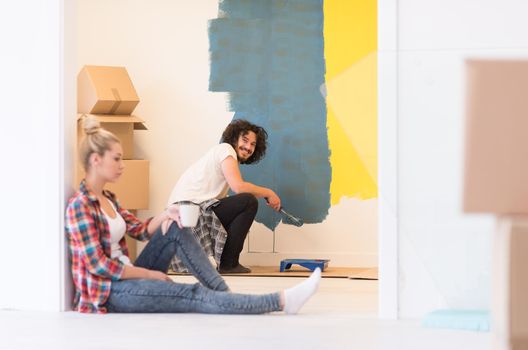 Happy couple doing home renovations, the man is painting the room and the woman is relaxing on the floor and drinking coffee