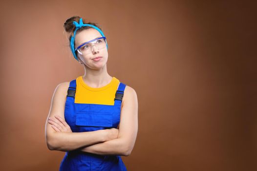 Strong fun young caucasian handyman woman in a yellow tank top and blue work overalls and safety glasses on a light brown background. A woman in a man's work, with her arms folded across her chest, is waiting and looking away. Copy space. Home renovation concept.