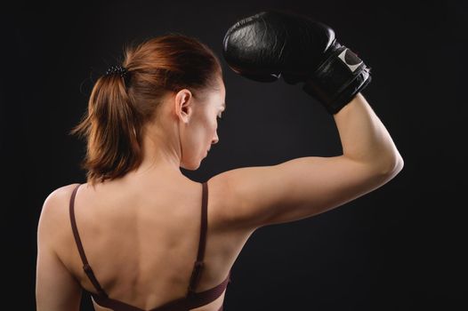 The back view of a Caucasian woman in sportswear and boxing gloves shows a gesture of strength. Demonstrates biceps. Studio shot on a black background. Woman fighter.
