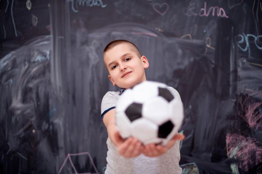 portrait of happy cute boy having fun holding a soccer ball while standing in front of black chalkboard