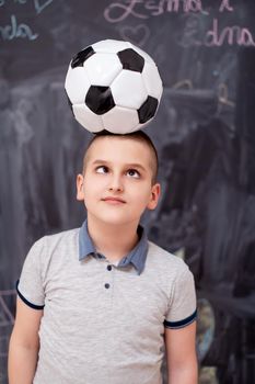 portrait of happy cute boy having fun holding a soccer ball on his head while standing in front of black chalkboard