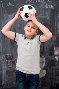 portrait of happy cute boy having fun holding a soccer ball on his head while standing in front of black chalkboard