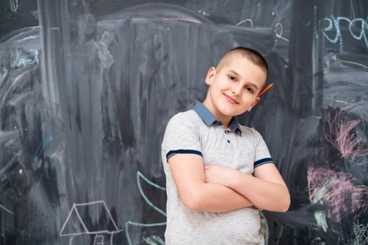 portrait of happy boy with orange wooden pen behind the ear standing in front of black chalkboard