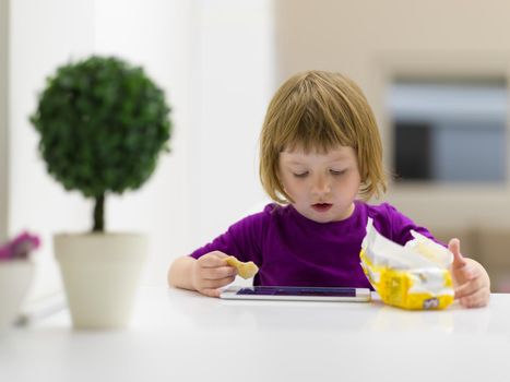 cute little girl eating a cookie while playing games on tablet computer at home