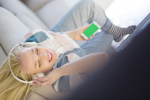 Young beautiful girl enjoying music through headphones, laying on sofa at home