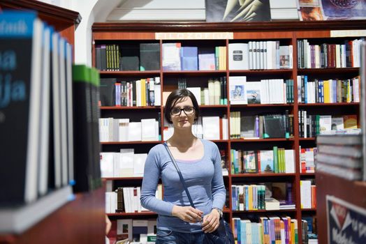 Pretty female student standing at bookshelf in university library store shop  searching for a book