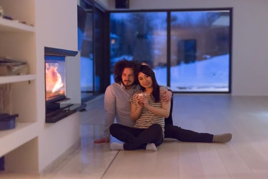 Young romantic multiethnic couple sitting on the floor in front of fireplace at home, talking and drinking tea on cold winter night