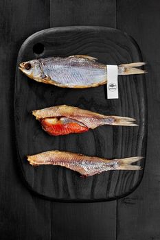 Whole salted air-dried roach fish with label on tail and two peeled fish with caviar served on black wooden board. Popular savory snack