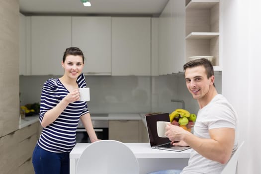 happy young couple with laptop computer enjoying morning coffee in modern kitchen at home