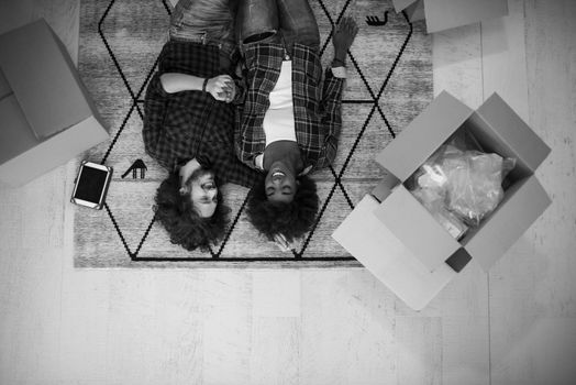 Top view of attractive young multiethnic couple moving, holding hands, looking at camera and smiling while lying among cardboard boxes