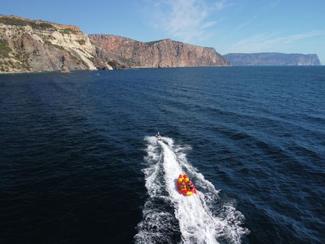 Happy people swim on air mattress behind a high speed water bike. Tourists ride on inflatable watercraft boat. Sea attraction. Water boat skating people on large Inflatable raft. Aerial copter view