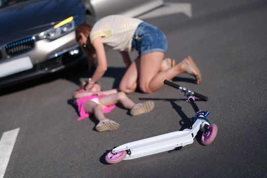 Little girl lying on the asphalt with scooter and her mother after collision with car. Baby pedestrian hitting concept