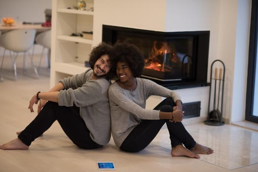 beautiful young multiethnic couple with tablet computer sitting on the floor of their luxury home in front of fireplace at autumn day