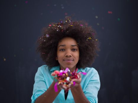 beautiful young black woman celebrating new year and chrismas party while blowing confetti decorations to camera isolated over gray background