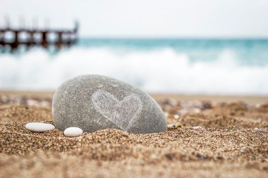 A round stone with a heart symbol lies on the golden sand of the beach on a blurred background of the sea