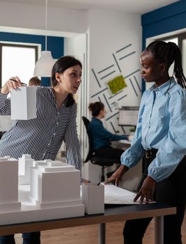 Architect presenting foam scale building model to engineer coworker standing next to residential project maquette in modern architectural office. Team of architects doing creative thinking.