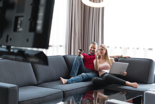 Young couple relaxes on the sofa in the luxury living room, using a laptop and remote control